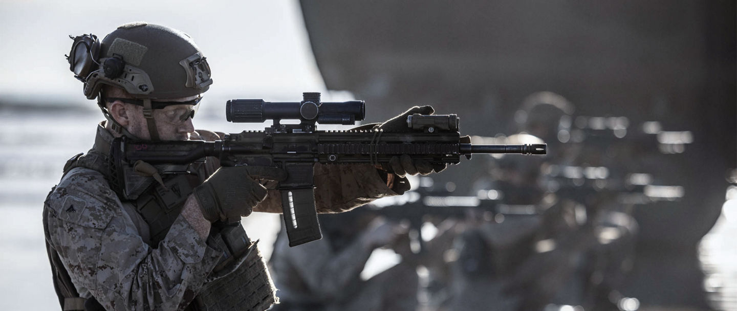 A Marine, in profile, looks down the scope of his rifle. He is on a beach with other Marines. They just got off an amphibious vehicle. 