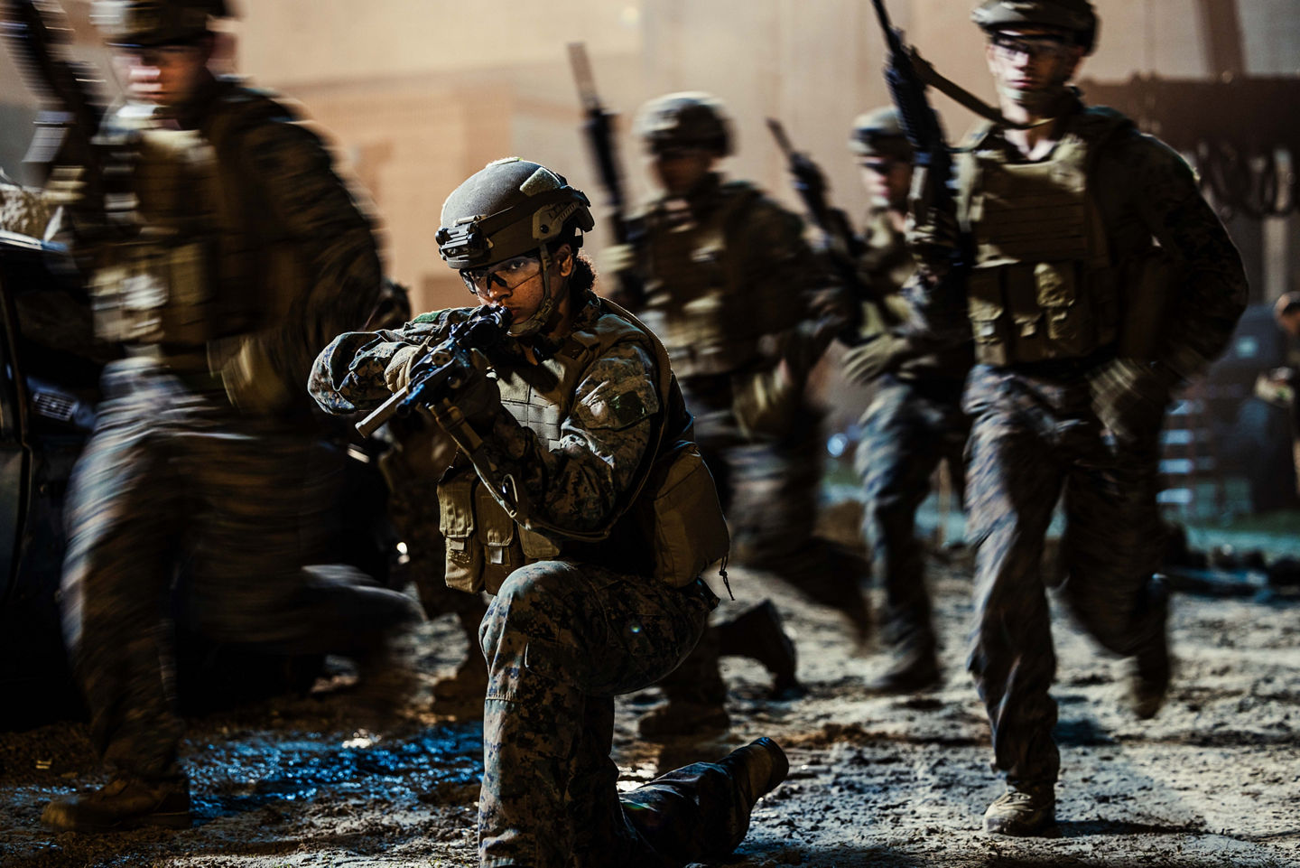 A Marine takes a knee and aims a rifle while fellow Marines in full combat gear advance around them during a low-light operation.