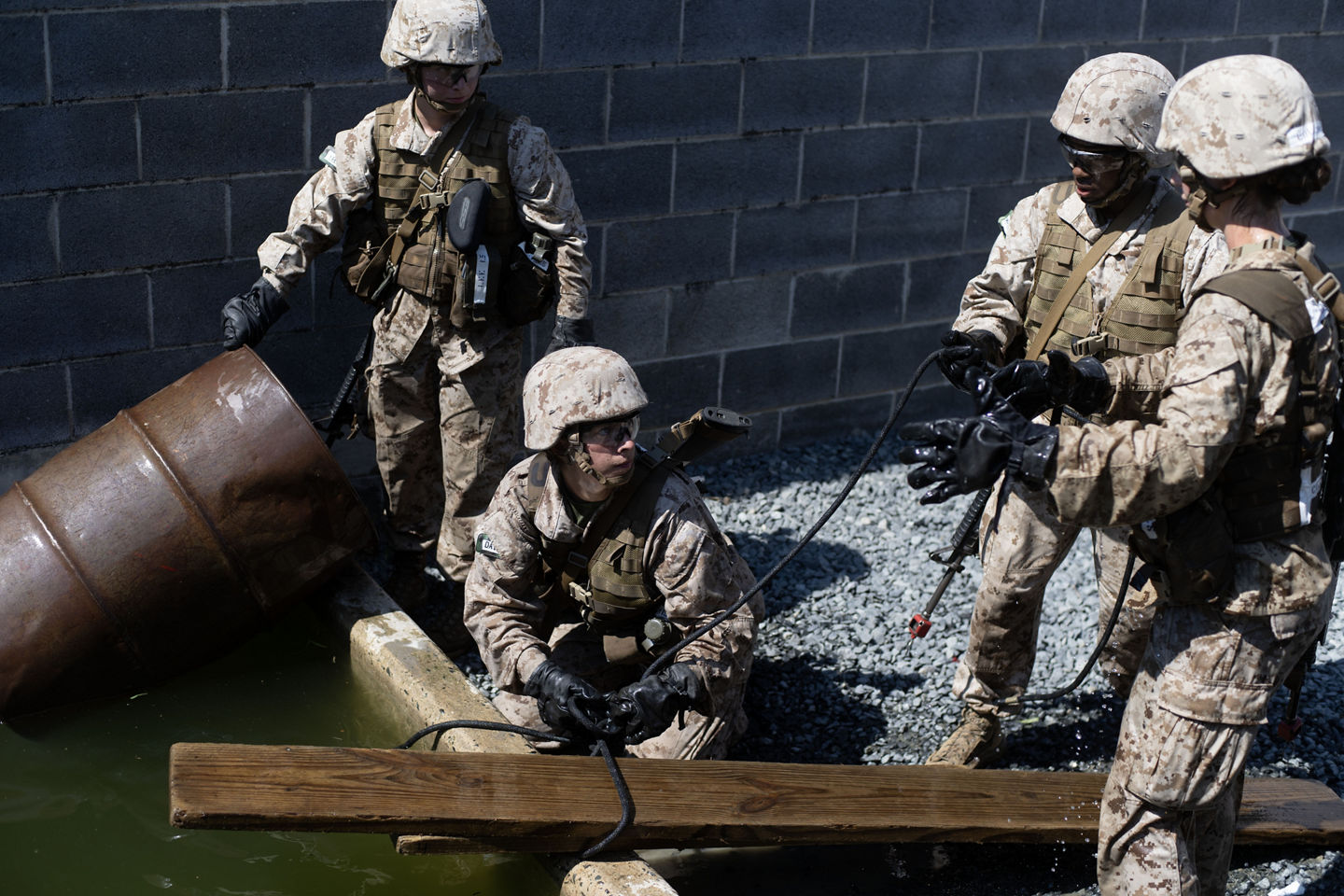 Marine officer candidates in camouflage utilities and helmets work together to move a rope across a barrel-and-plank water obstacle at the Leadership Reaction Course.