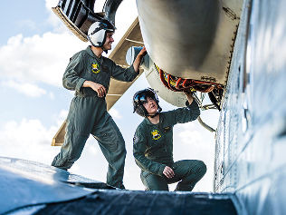 Two Marines in flight suits and helmets examine electrical wiring on the engine of a helicopter.