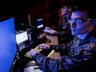 Two Marines work at computers in a command center. They sit in the dark in front of numerous monitors.