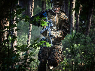 A Marine in a camouflage uniform sprints quickly through the woods. He holds his rifle in both of his hands.