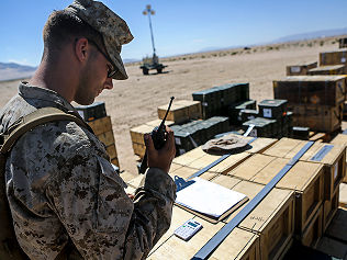 A logistics Marine talks on the radio as he oversees the movement of supplies stacked on pallets.