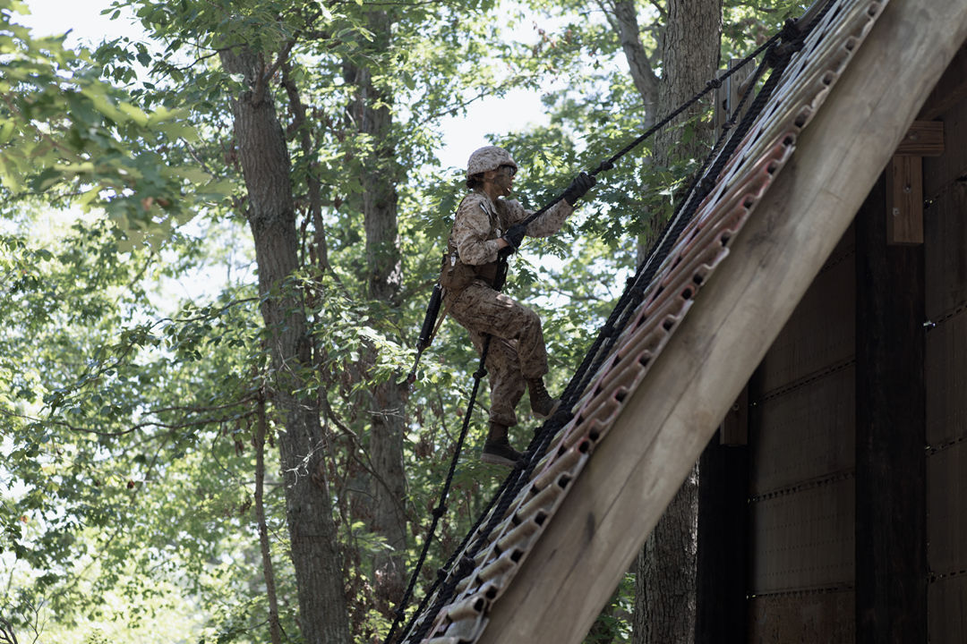 A Marine uses a rope to climb up a steeply angled wall. They are completing a combat field course.
