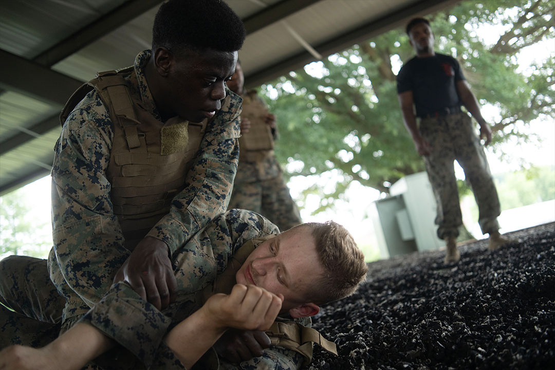 Two Marine recruits wrestle on the ground. One pins the other. A drill instructor oversees the training.