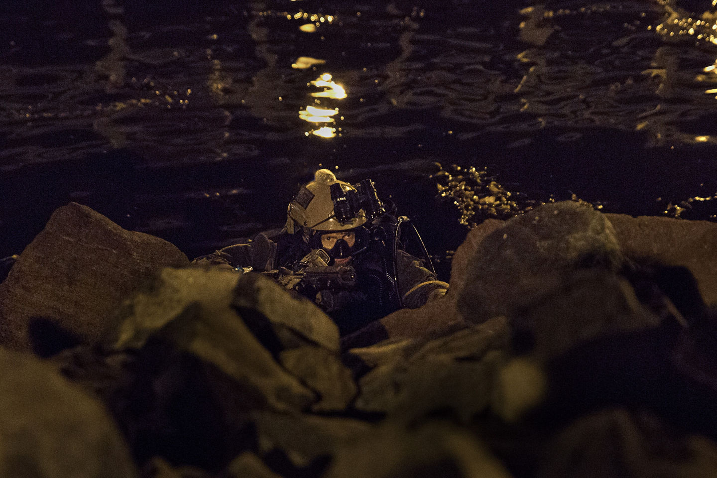 A special operations Marine in goggles and scuba gear rises out of the water at night. 