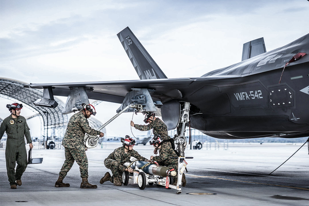 Marines in camo and flight suits load a missile onto the wing of a F-35B on an airfield tarmac. 