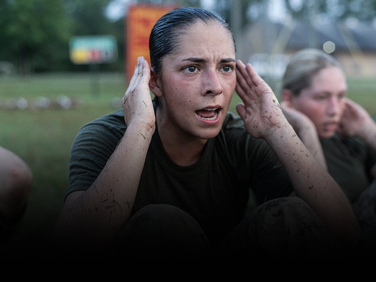 Female Marine recruit performs physical training during enlistment preparation with other recruits in background.