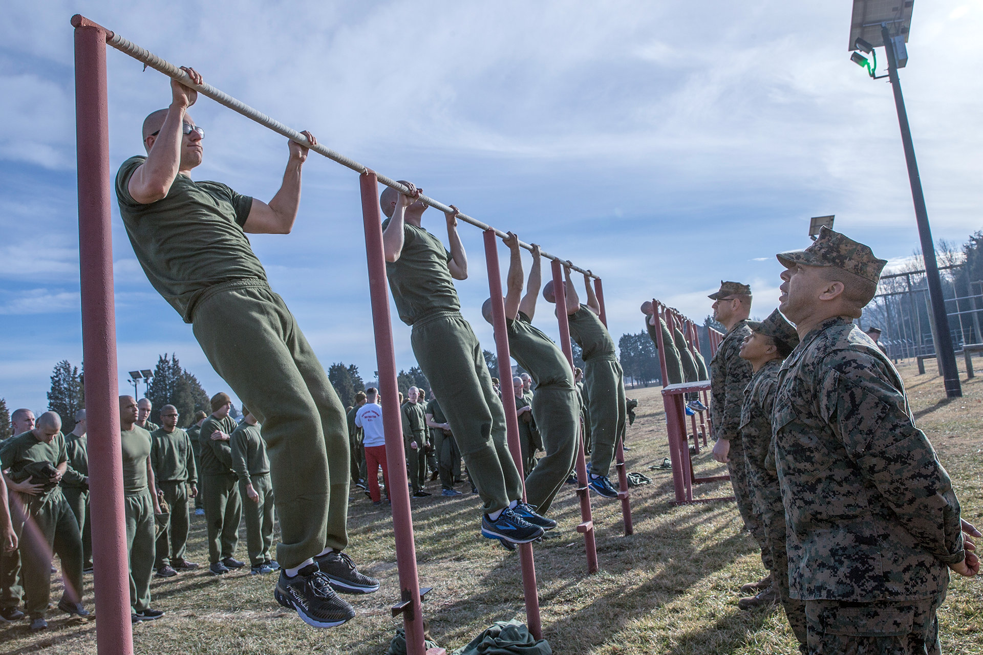 Marine recruits performing pullups on bar while instructors observe during physical fitness assessment.