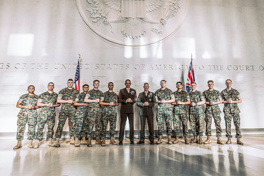 Marines in woodland camouflage uniforms with senior officers standing ceremonially at embassy under American flag and presidential seal.