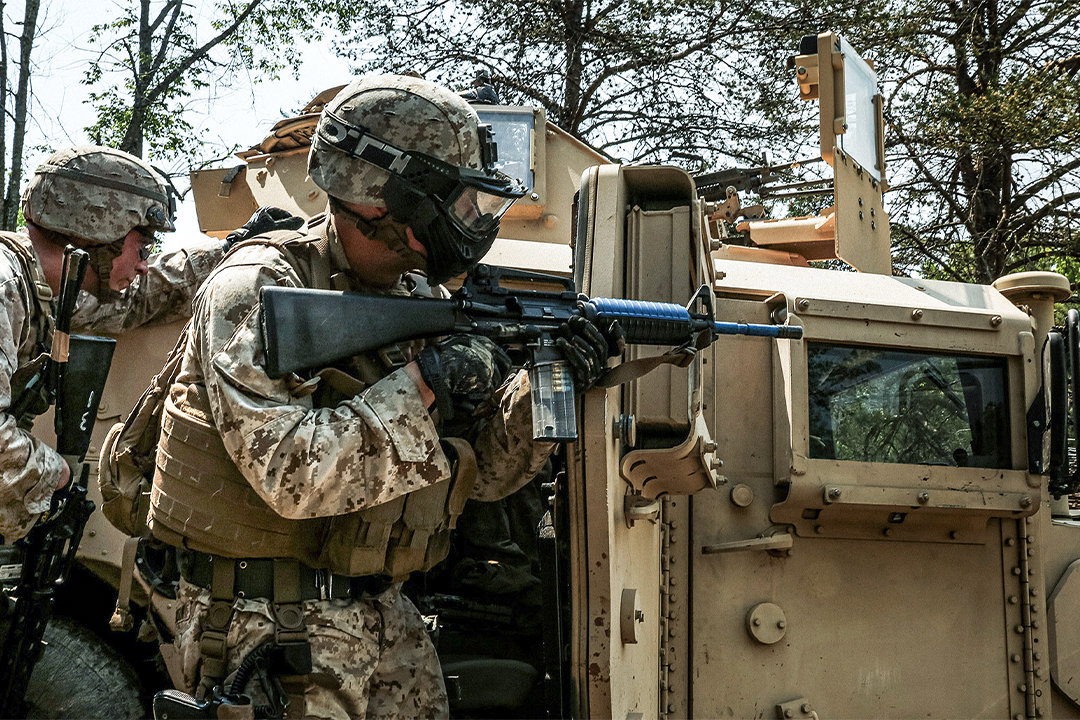 Marines in desert camouflage with rifles conducting tactical training exercise beside armored military vehicle.
