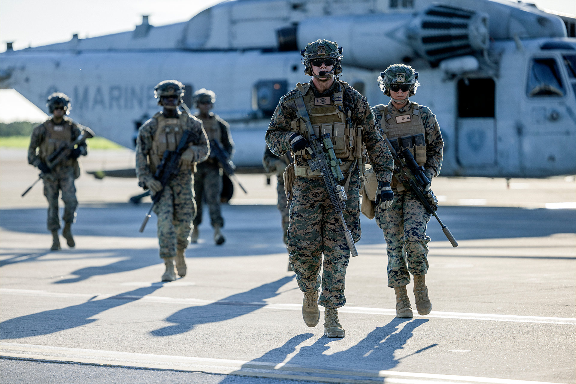 Marines in woodland camouflage with tactical gear and rifles walking across aircraft carrier flight deck during deployment operations.