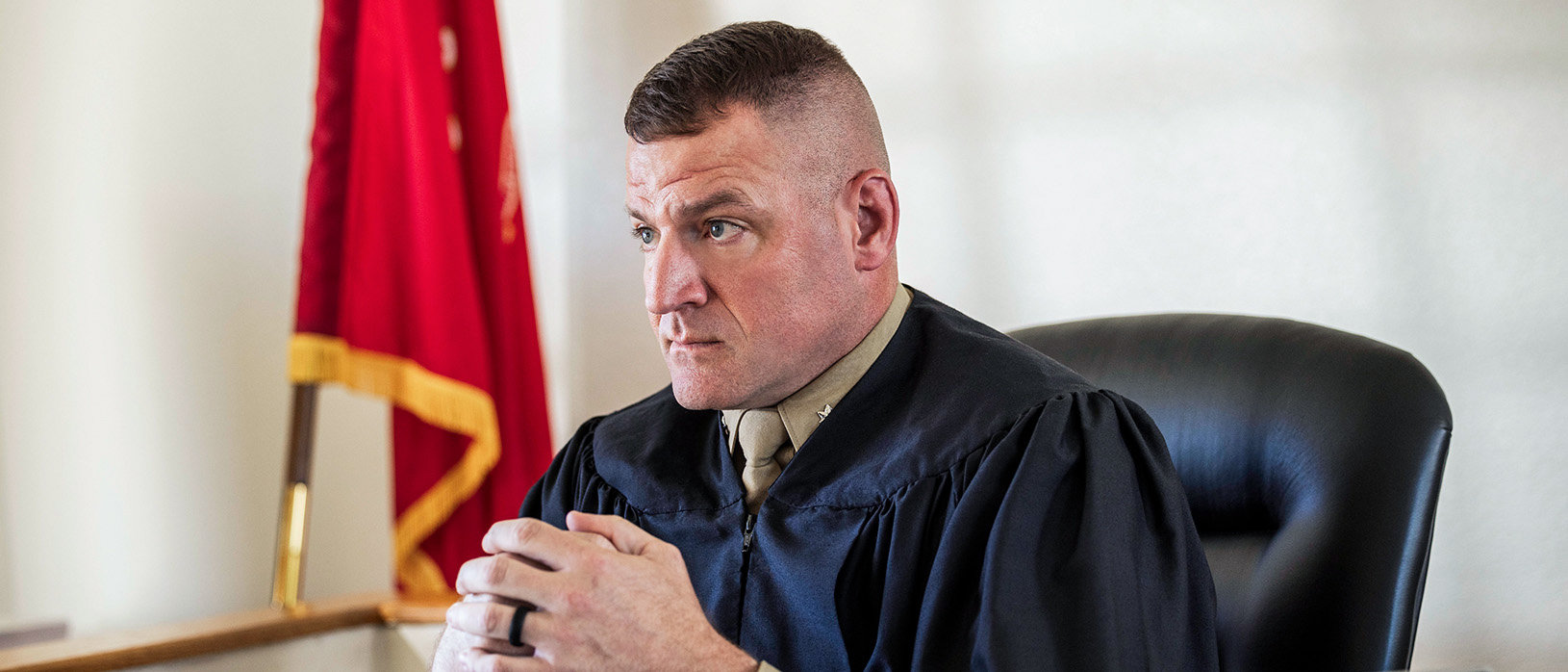 Marine judge advocate in judicial robes seated at desk with American flag during legal proceedings.