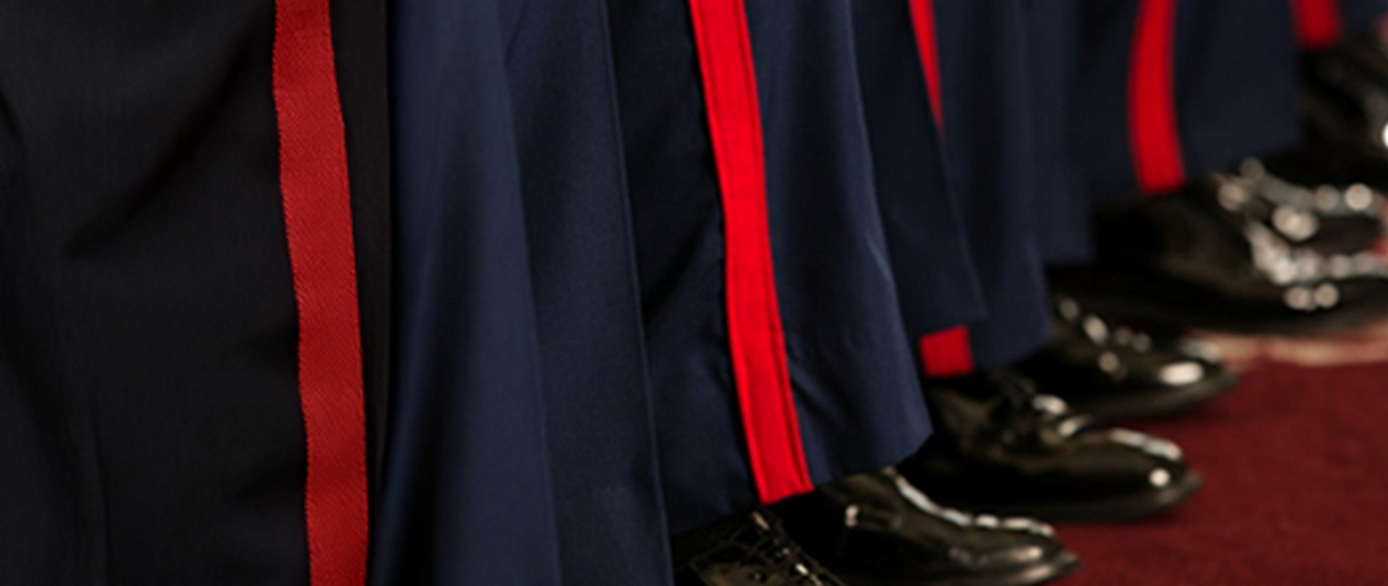 Close-up of several Marines in dark blue uniforms with red stripes and shiny black shoes, standing on a red carpet.