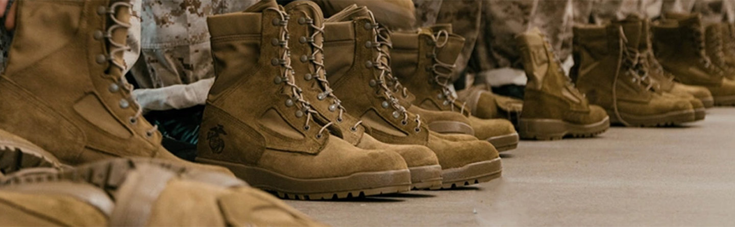 Multiple pairs of tan Marine combat boots with laces, one featuring the Marine Corps emblem, lined up on a dark surface.