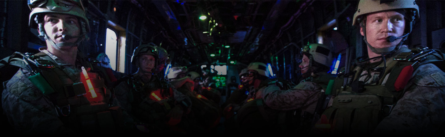 Several Marines in combat military gear sit inside a dark transport helicopter, illuminated by red and green lights.