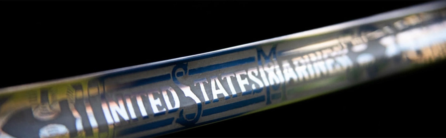 Close-up of a metallic, Mameluke sword with "UNITED STATES MARINES" engraved on it, against a dark background.