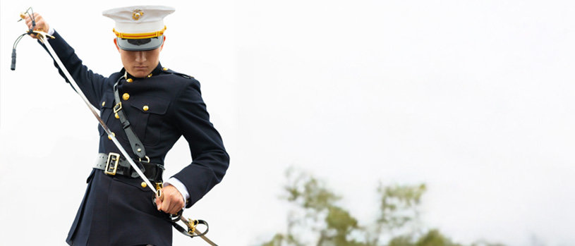 A Marine in dress uniform sheathing a mameluke sword, looking down, with a blurry outdoor background.