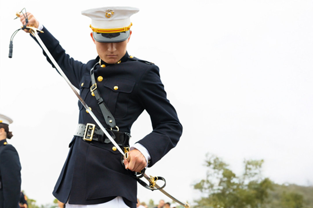 A Marine in dress uniform sheathing a mameluke sword, looking down, with a blurry outdoor background.