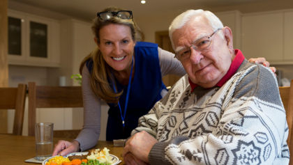 An older man being served a meal by a woman in a blue vest with a name tag on a lanyard 