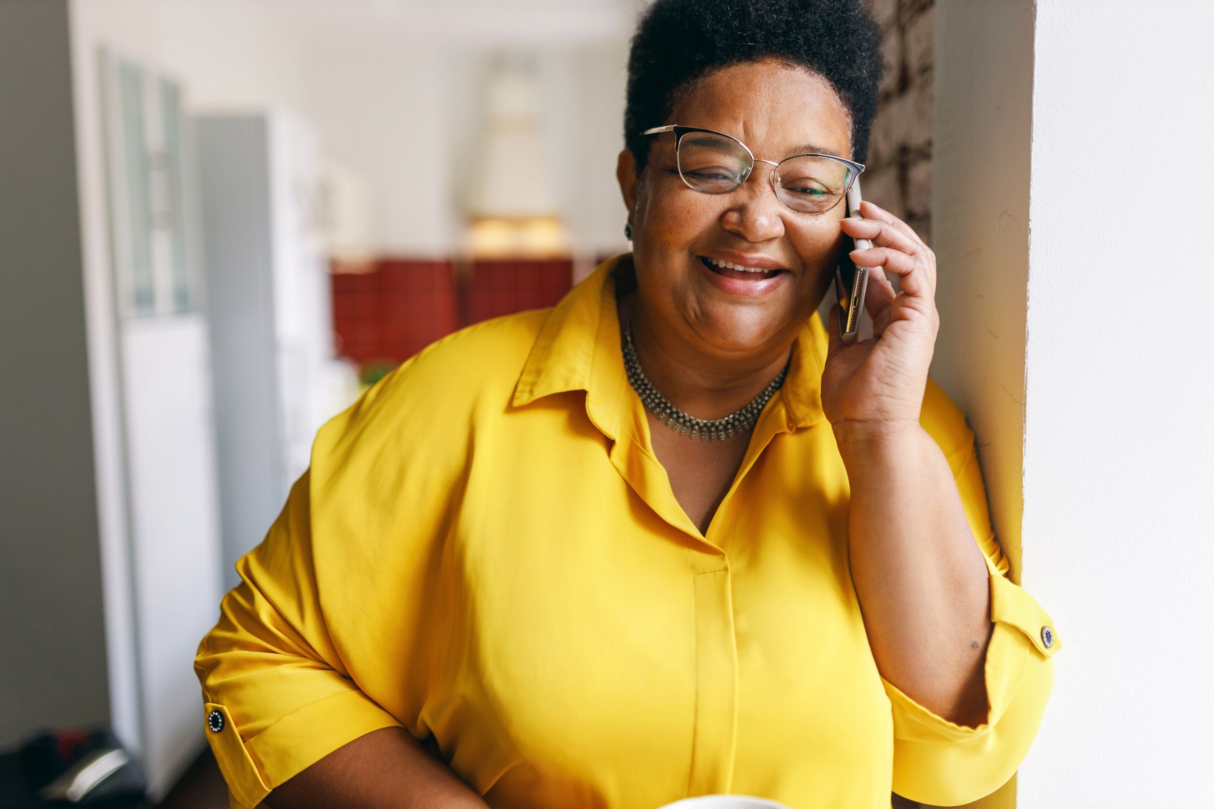 Happy smiling senior black plus size female in yellow shirt and glasses having nice conversation on phone with her best friend, smiling and laughing at jokes, sharing news and rumors