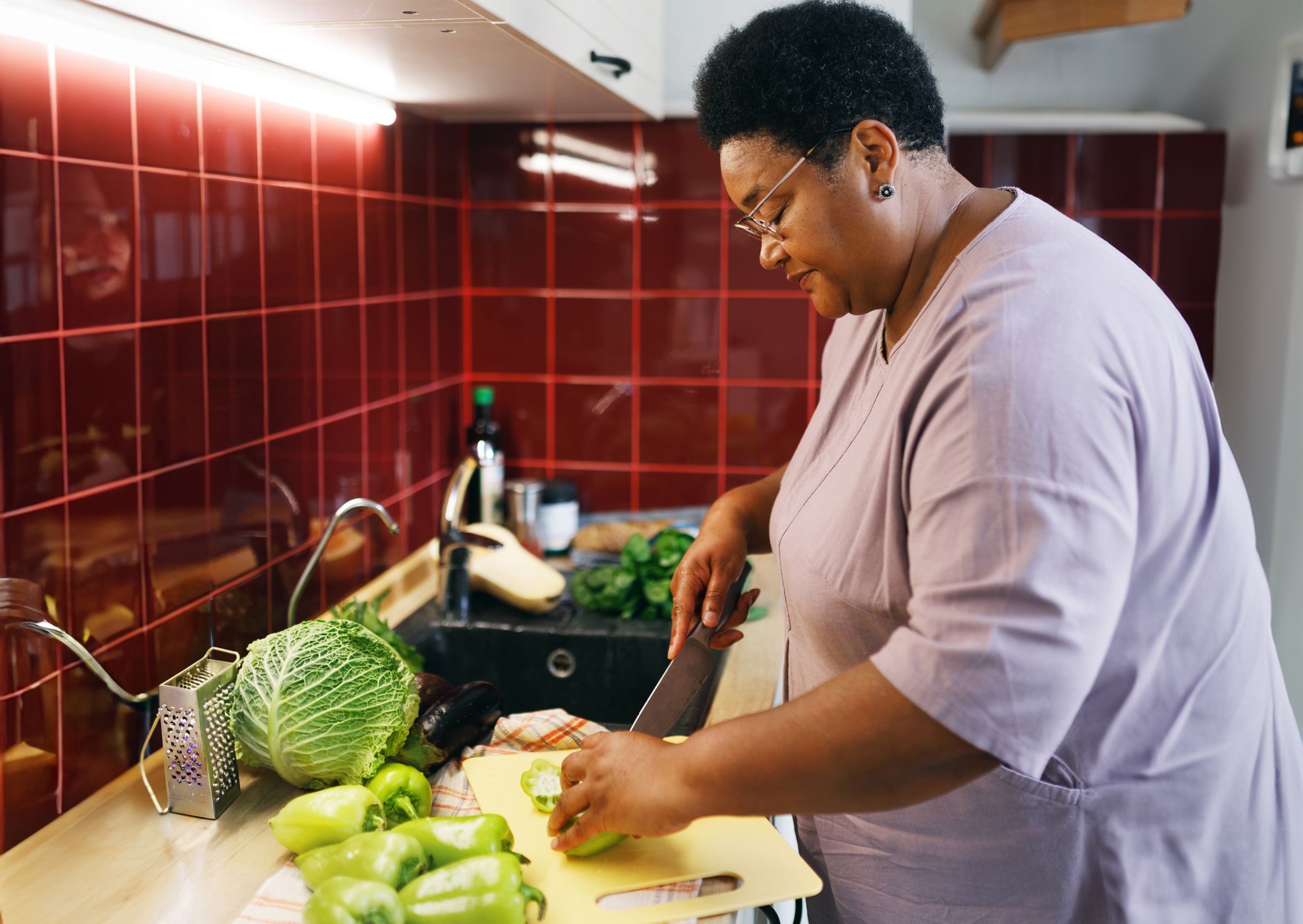 Side view of overweight african american senior woman cooking healthy veggie dinner standing next to counter chopping and slicing vegetables, zucchini and bell pepper, using new recipe