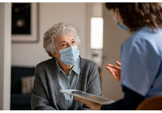 An elderly woman wearing face mask is discussing with a clinician who is holding a tablet