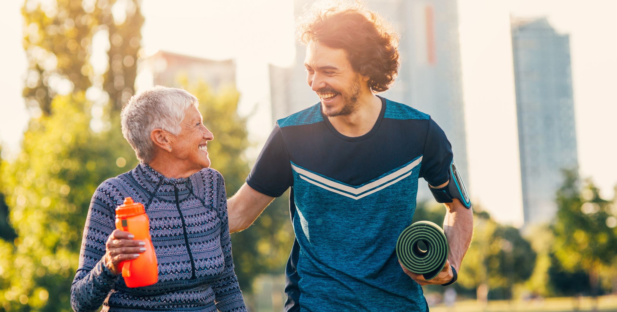 Young man with older woman, man holding yoga mat and smiling, woman holding water bottle and looking at him smiling. 