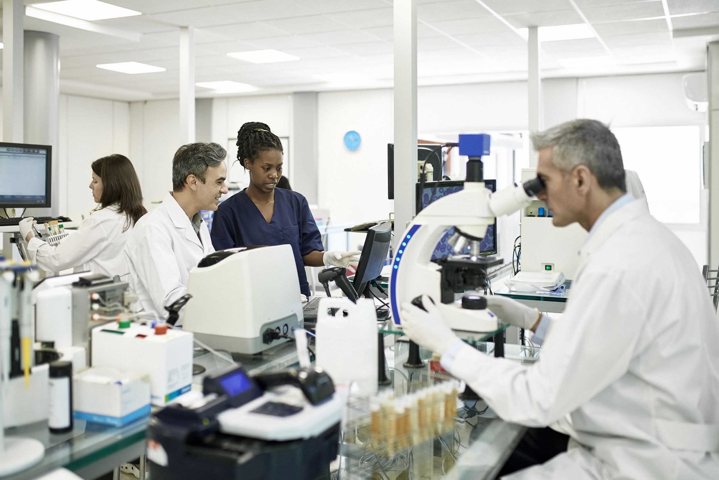 male and female pathologists in white coats with technician using microscope and studying data on computer in Clinical Analysis Laboratory.