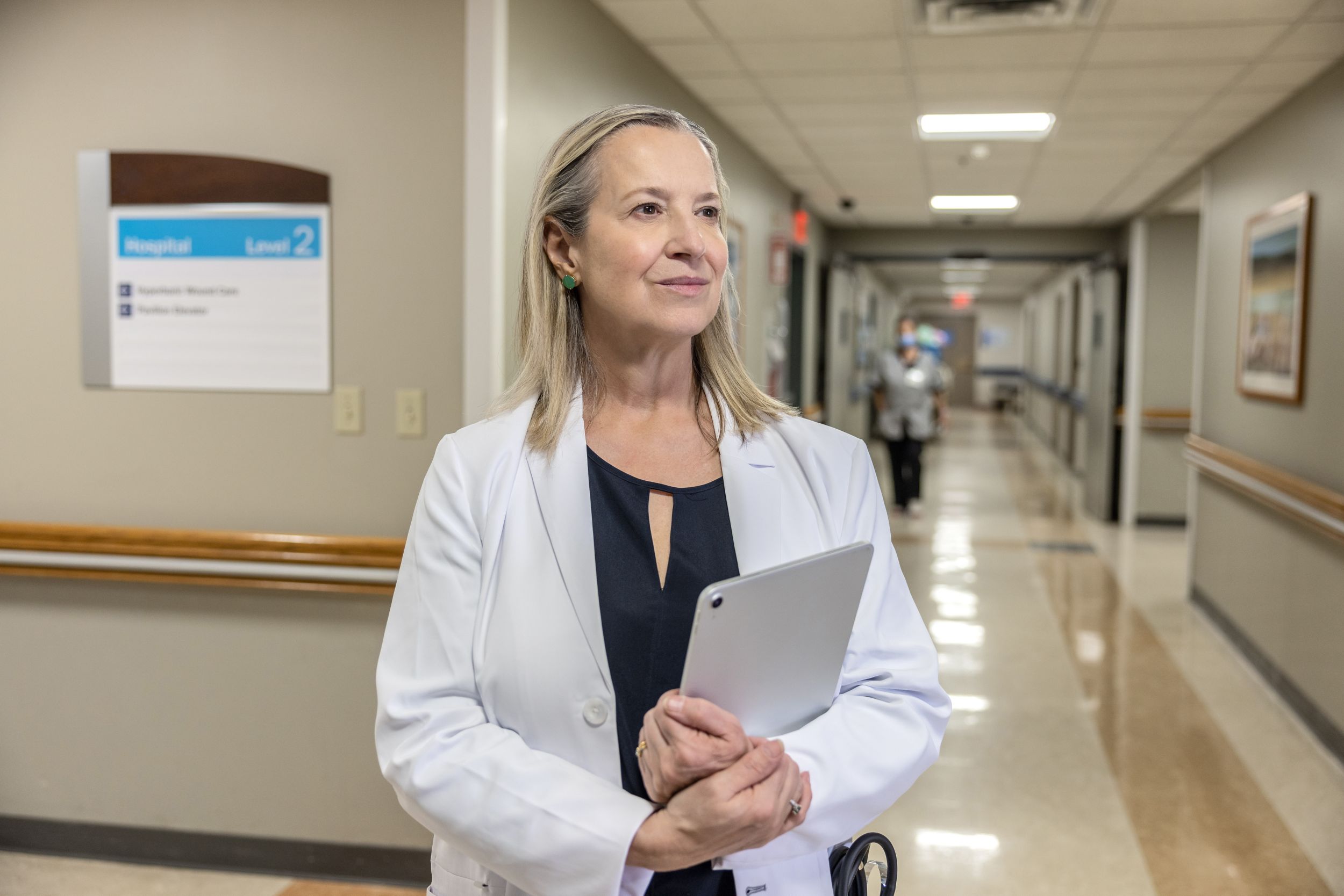 Female physician holding clipboard and standing in hallway of a medical facility.