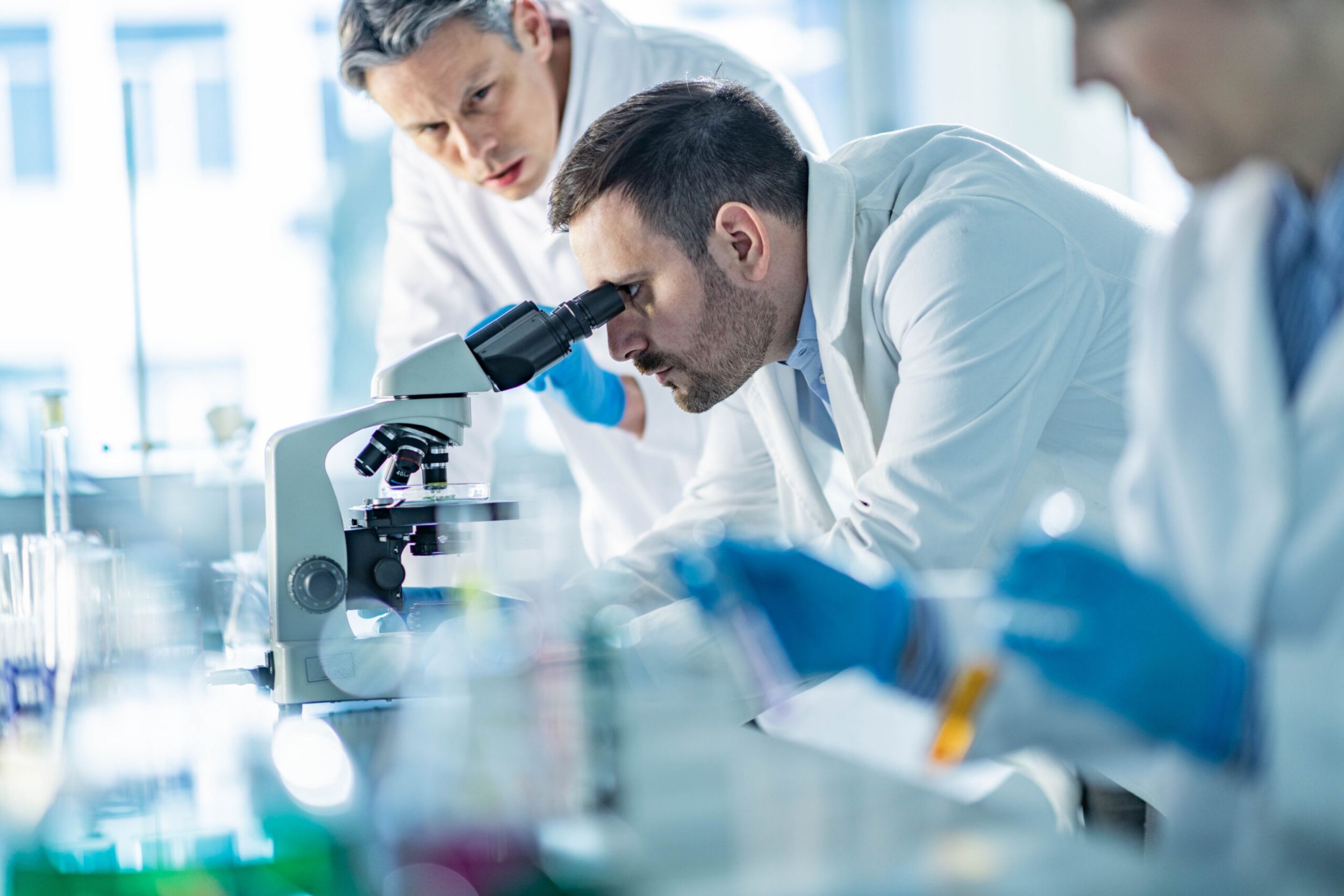 Young male chemist looking through a microscope while working with his colleagues on antiviral drug in a laboratory.