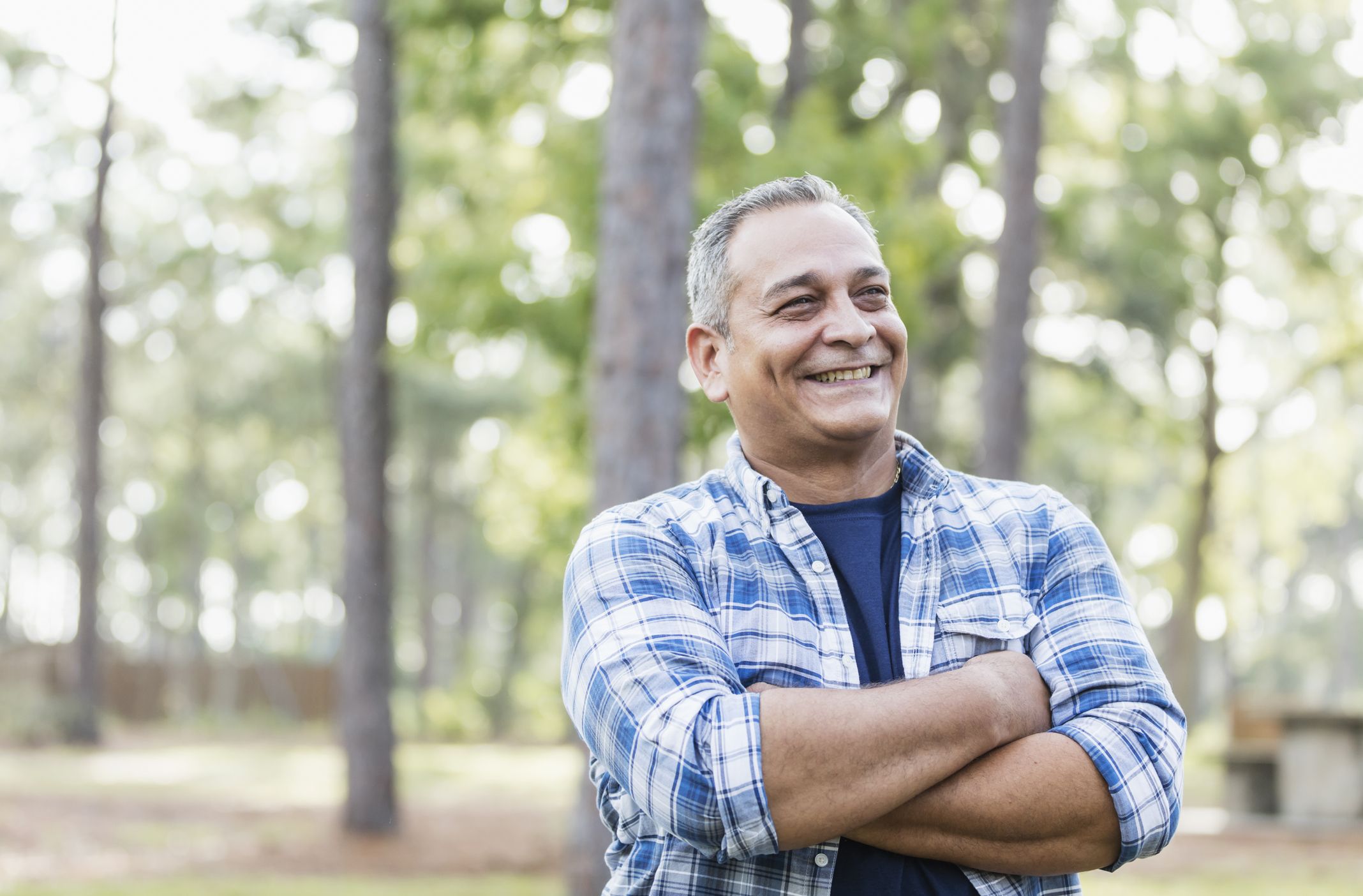 A mature Hispanic man in his 50s wearing a plaid shirt, standing in a park, smiling with his arms crossed.