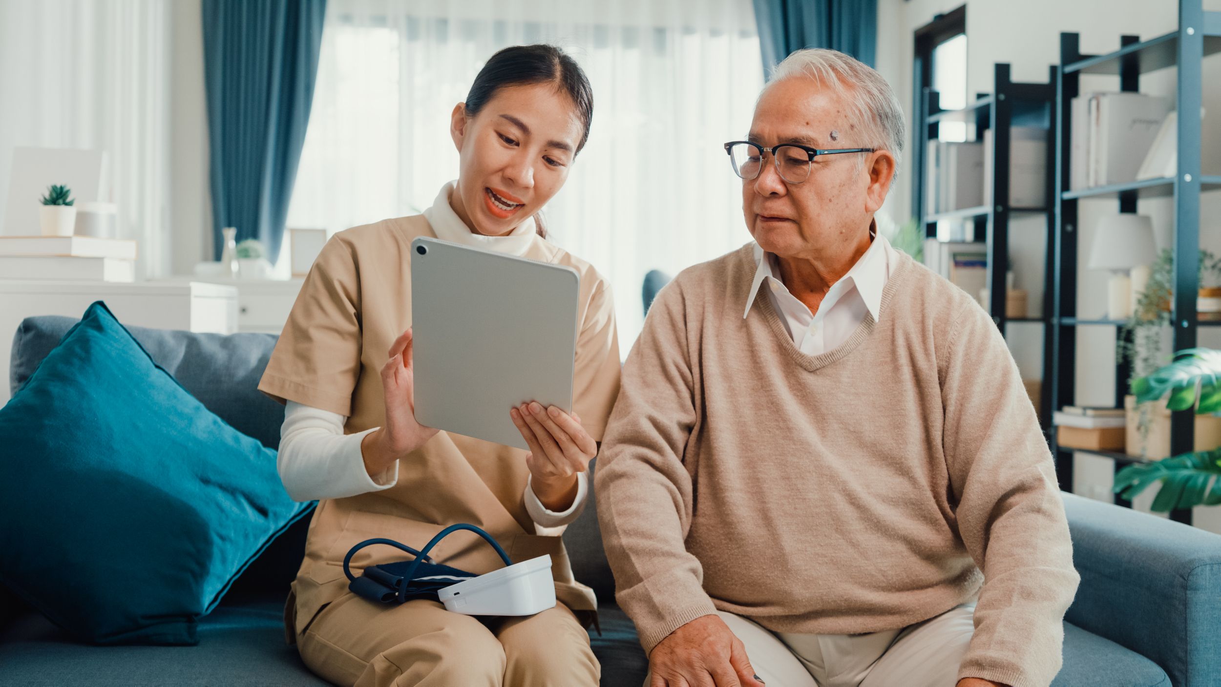 Young Asian female professional caregiver helps older Asian man use digital tablet on couch at home.