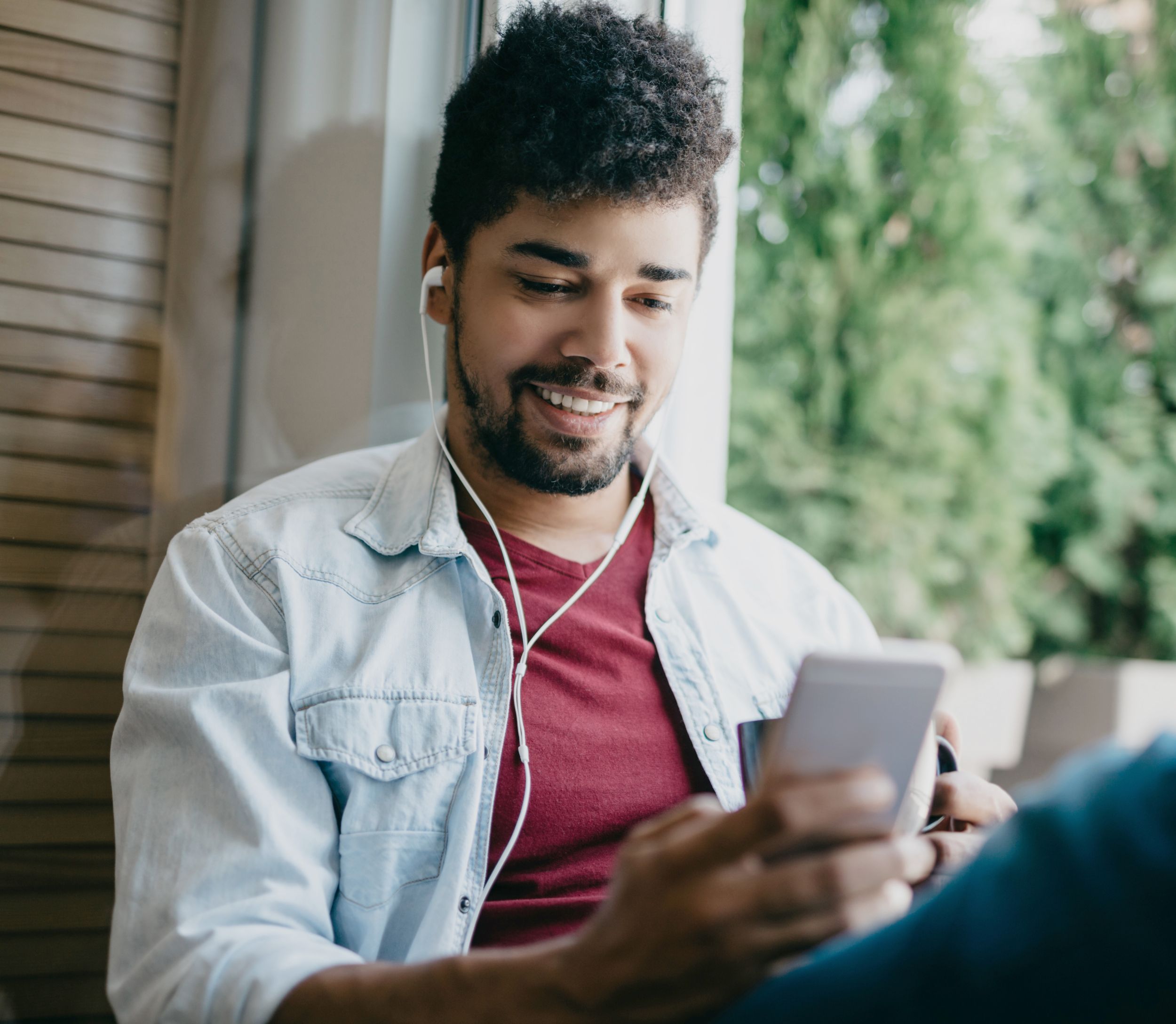 Young man with headphones sits against a window and looks at phone