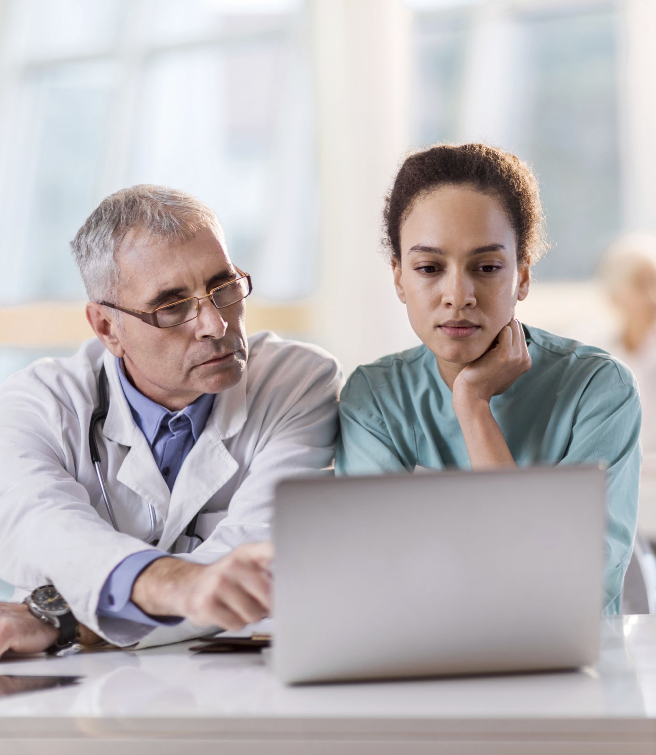 Doctor and a nurse sitting at doctor's office and using computer. There are people in the background.